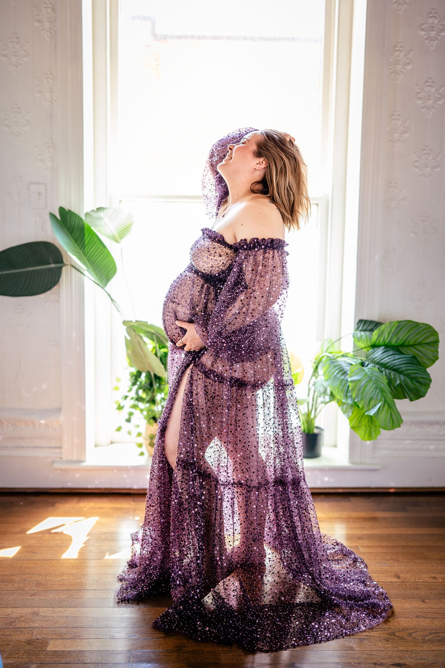 Pregnant woman in purple sparkly off-shoulder gown stands by sunlit window surrounded by tropical plants.