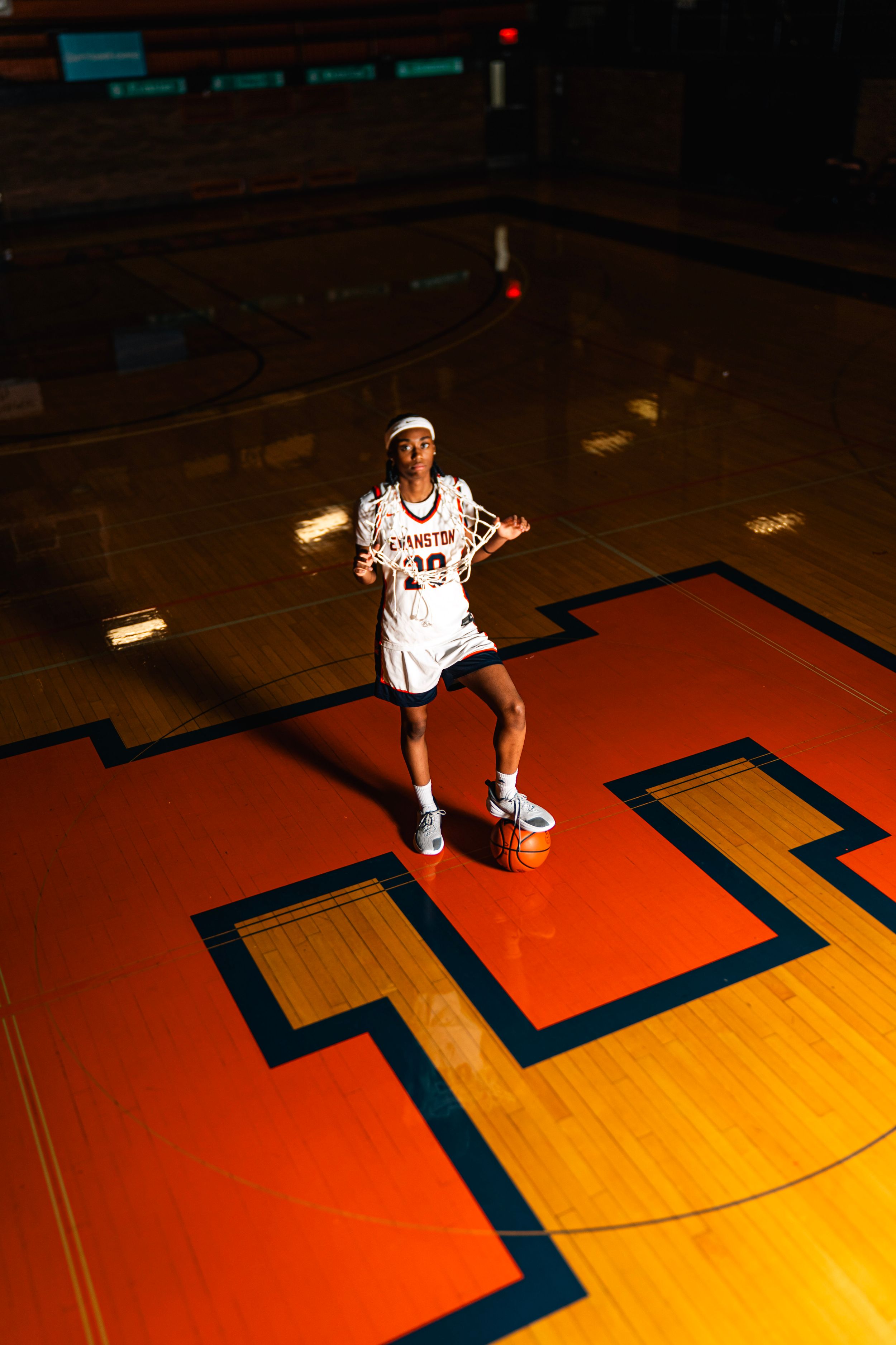 Basketball player in Evanston jersey standing on court with ball, dramatic overhead lighting on hardwood floor.