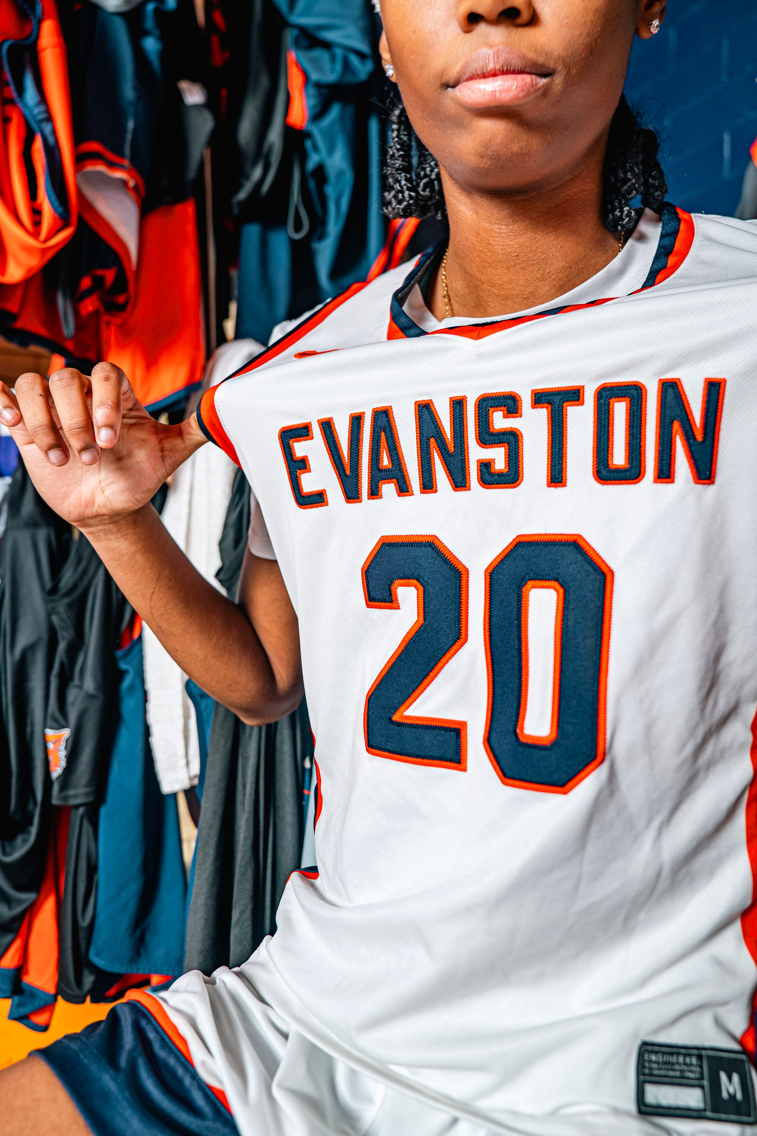Female athlete wearing Evanston #20 white basketball jersey poses in locker room surrounded by sports uniforms.
