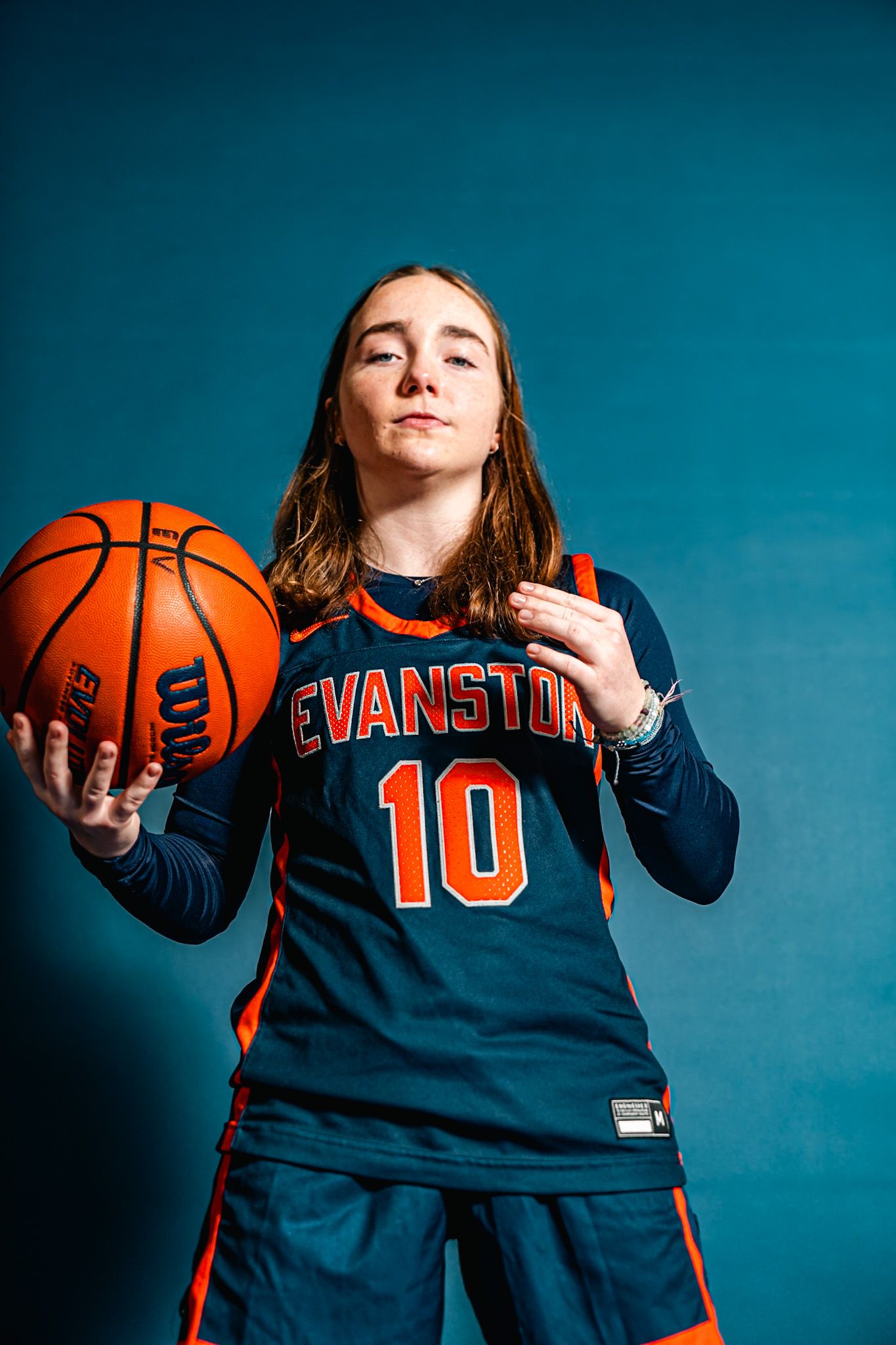 Evanston basketball player #10 poses with a Wilson ball in an orange and navy uniform against a teal background.