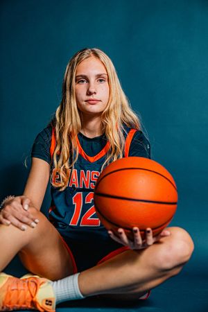 Young female basketball player #12 in Evans jersey poses with basketball on teal studio background.