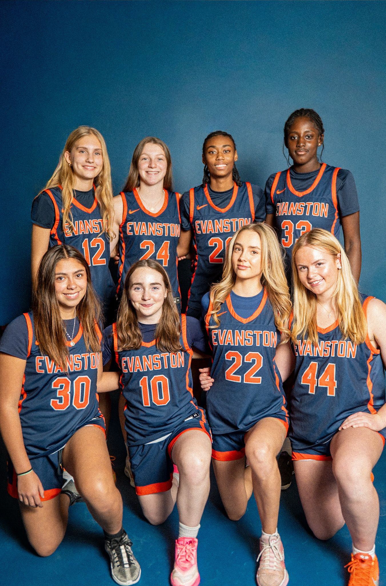 Eight Evanston girls basketball players pose for a team photo in navy and orange uniforms against a blue background.