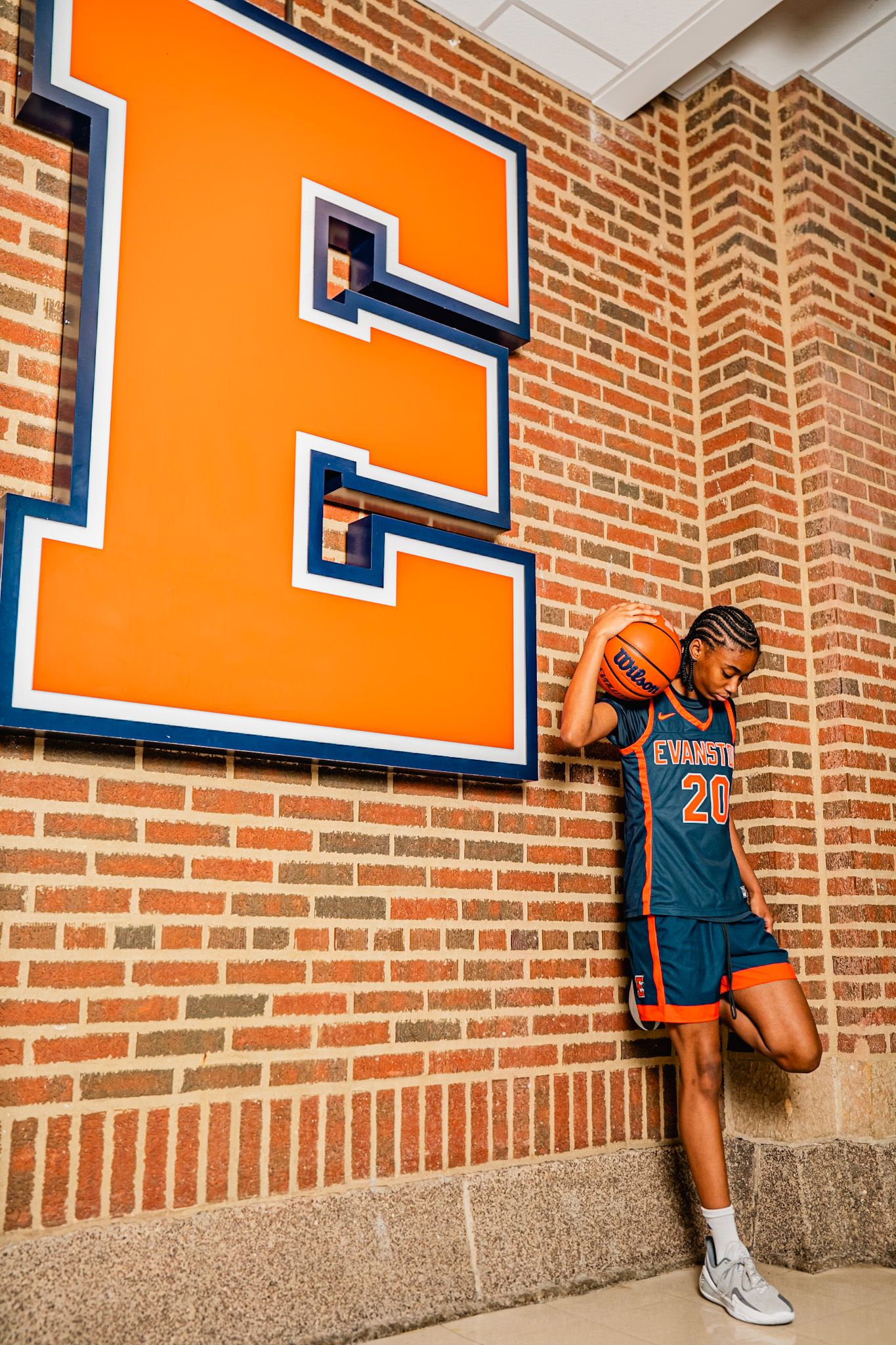 Basketball player in teal and orange Evansville jersey #20 poses with ball against brick wall with large E logo.