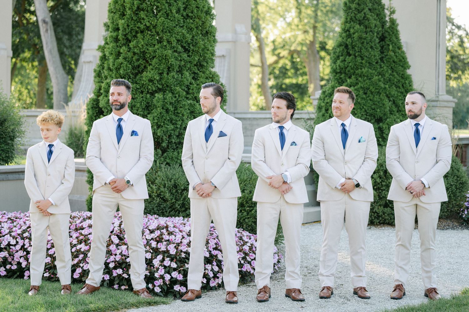 Armour House Wedding - Six groomsmen in tan suits with blue ties stand outdoors at a wedding venue surrounded by lush greenery.