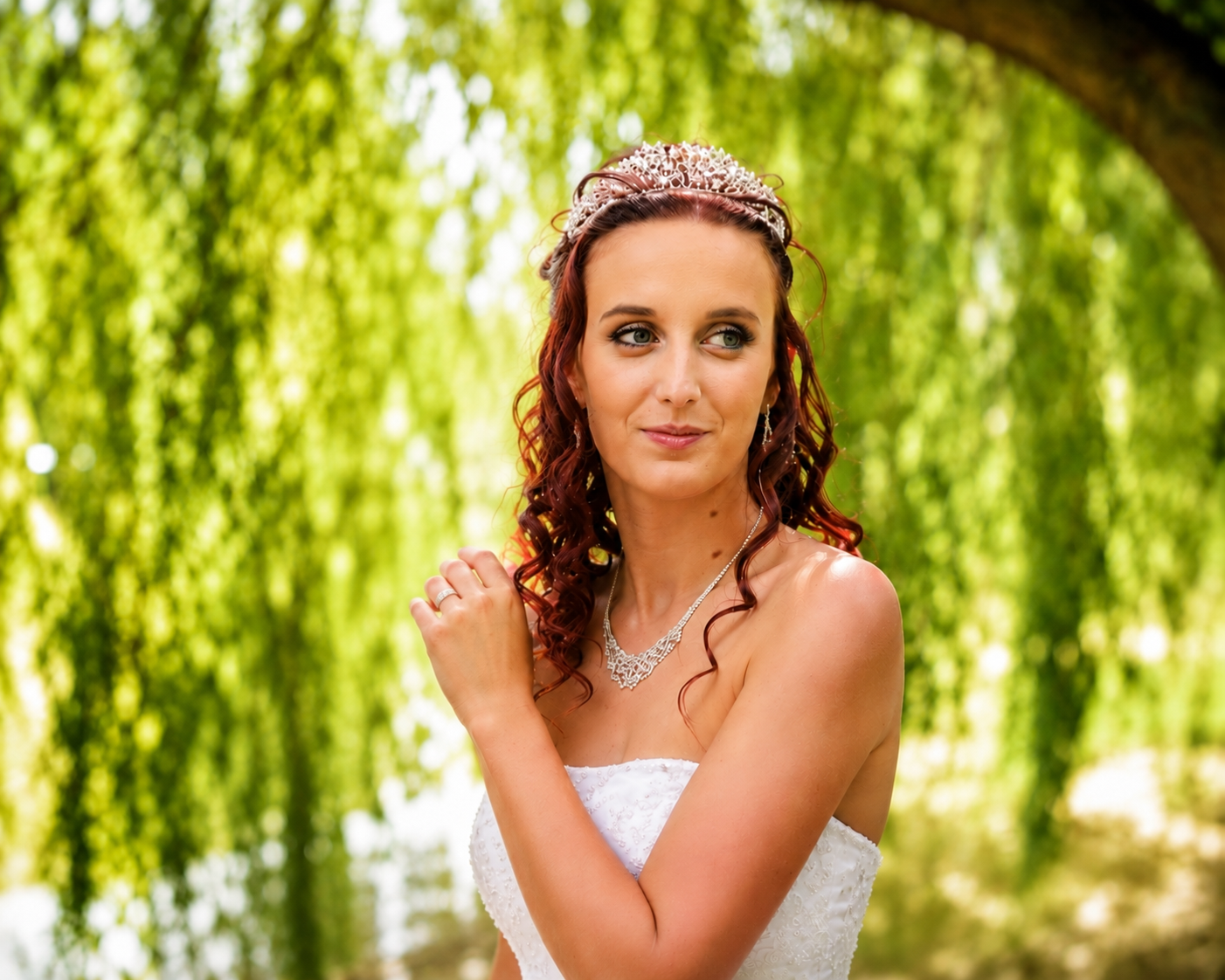 Beautiful bride wearing tiara and jewelry poses under weeping willow tree outdoors.