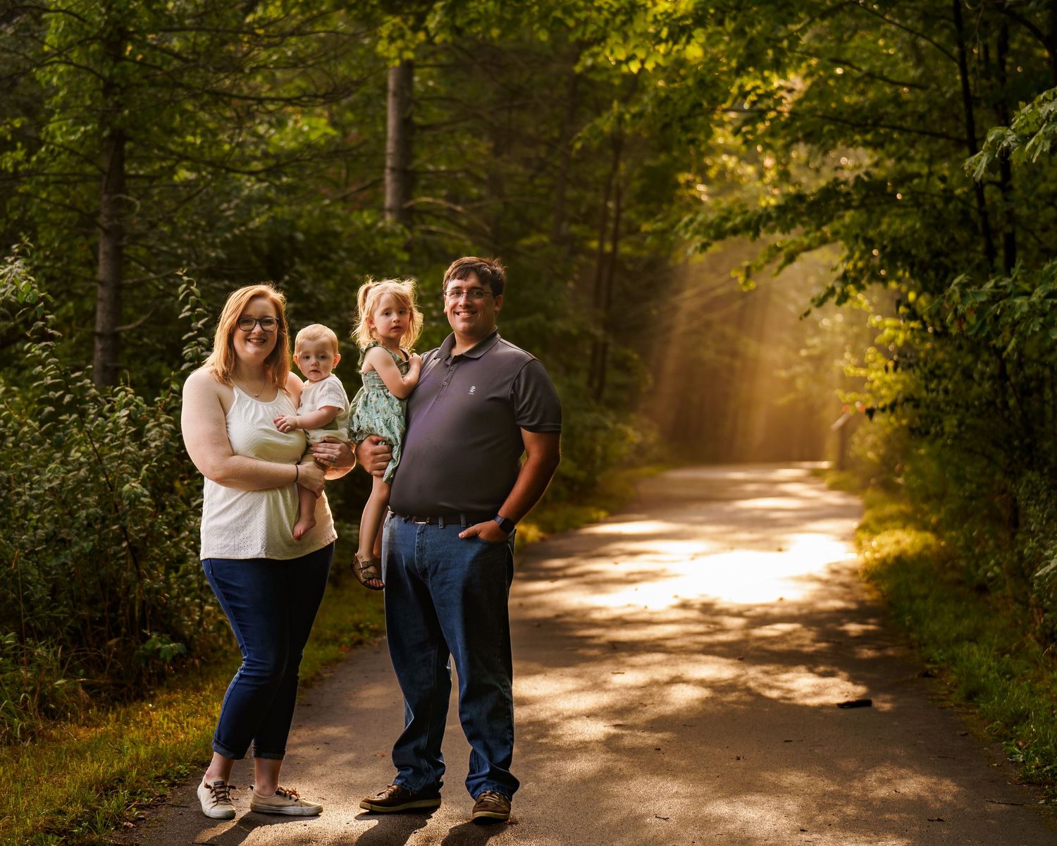 Family of four posing on a sunlit forest path surrounded by lush green trees during golden hour.