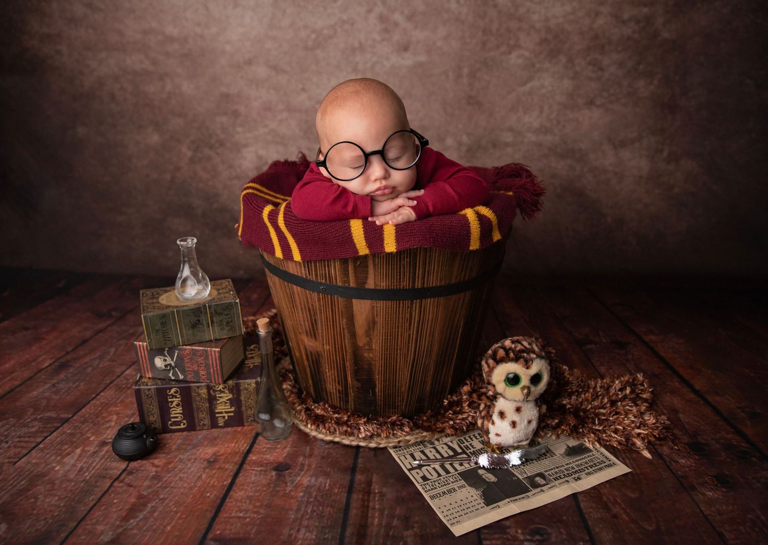Newborn baby dressed as Harry Potter in a wooden bucket with round glasses, Gryffindor scarf, owl, and books.