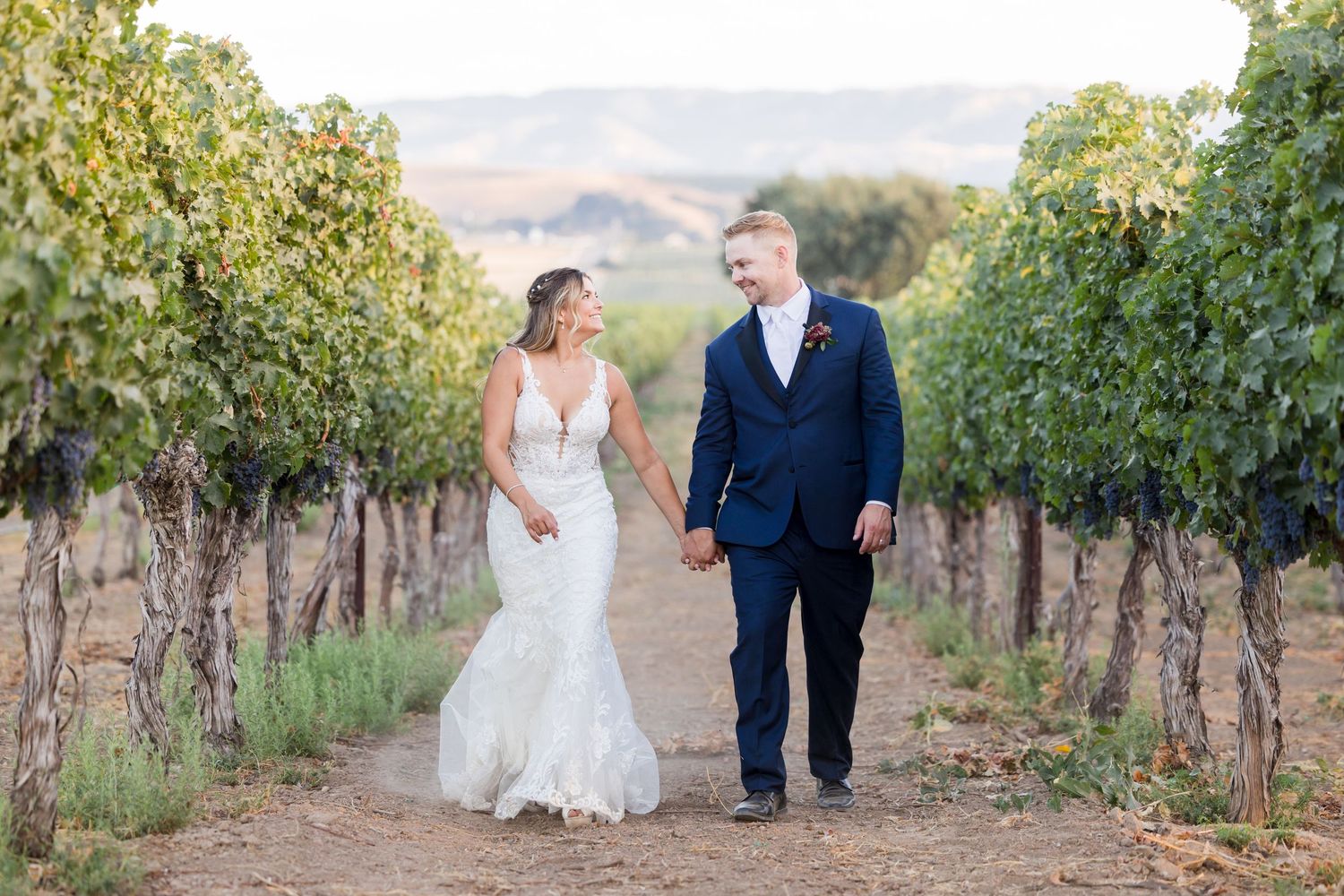 Black and white photo of a smiling bride in lace gown holding bouquet as groom in suit whispers in her ear.