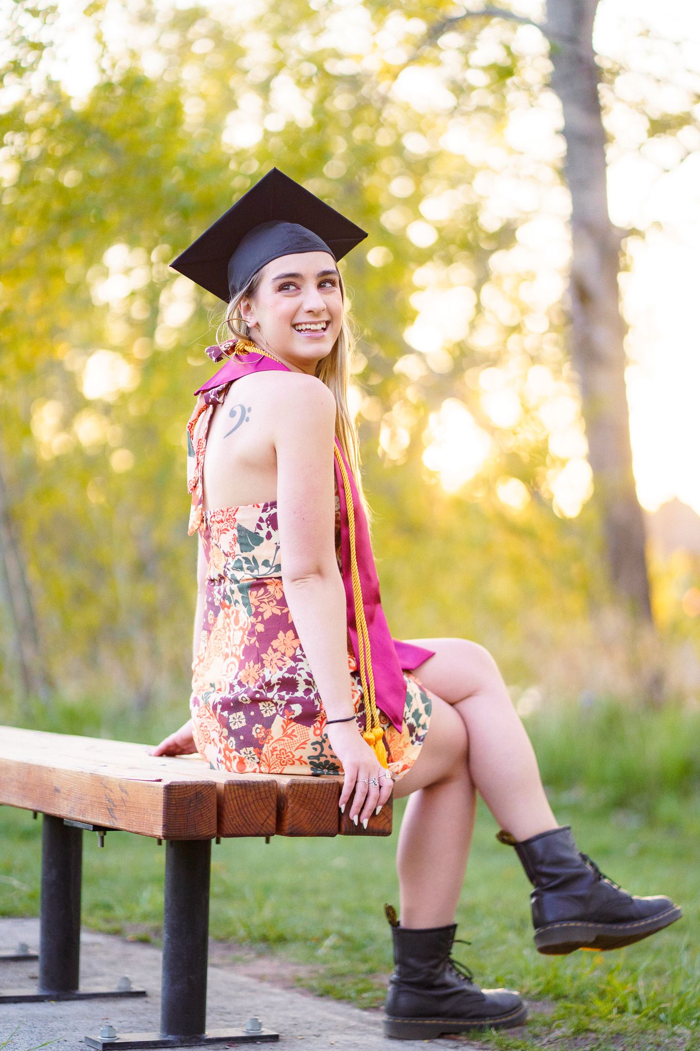 Smiling graduate in floral dress and cap sits on bench outdoors, wearing maroon sash and black boots.