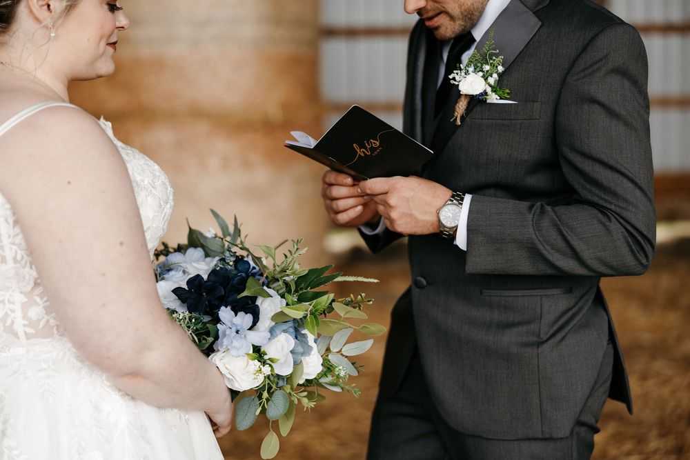 Groom reads vows from black book while bride holds blue and white floral bouquet during intimate barn wedding ceremony.
