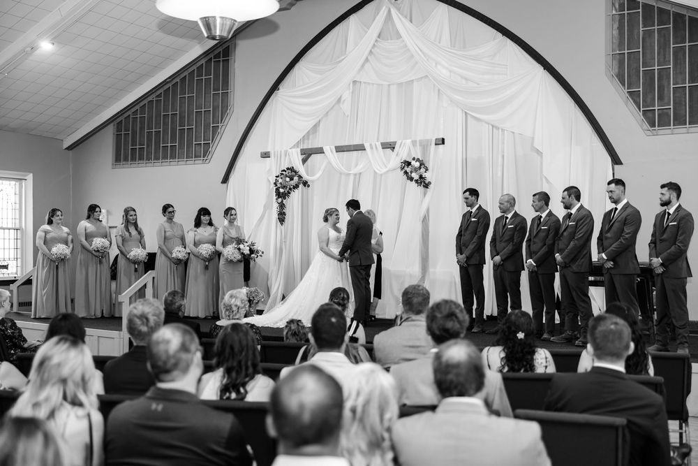Black and white photo of wedding ceremony in church with bride, groom, bridesmaids and groomsmen at decorated altar.