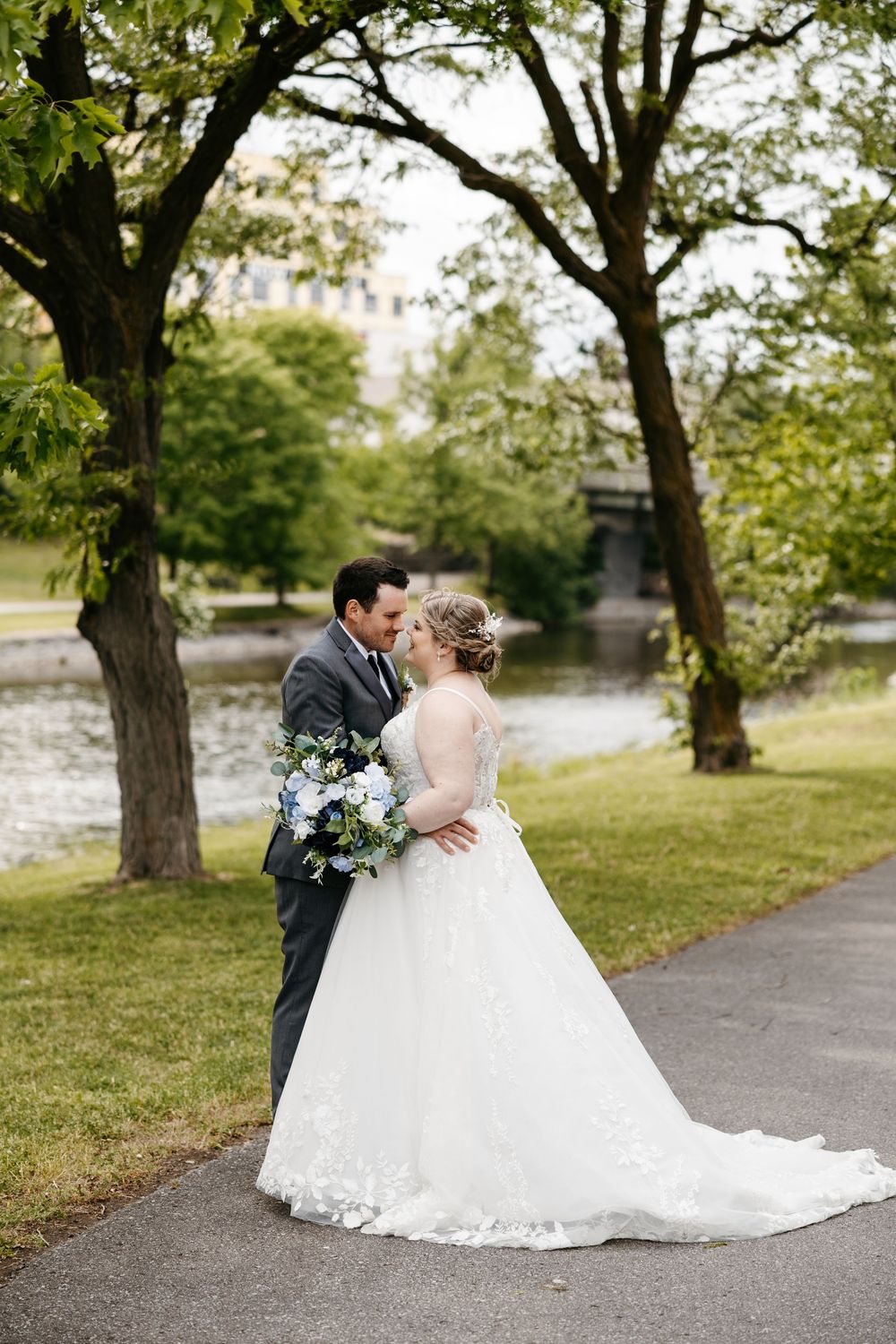 A bride in a white lace gown and groom in gray suit share a kiss near a serene pond surrounded by lush green trees.
