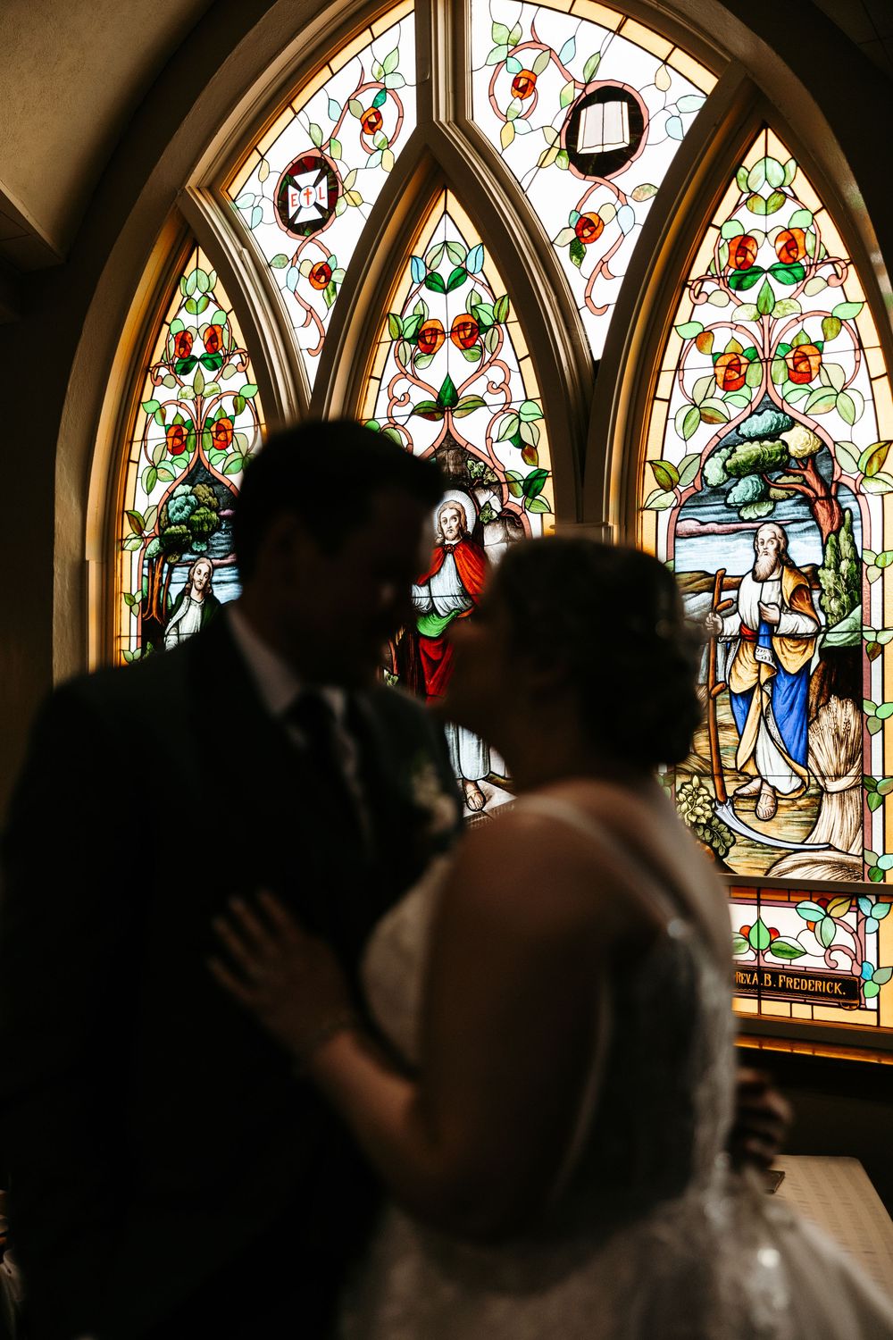 Silhouette of bride and groom sharing a romantic moment before a stunning stained glass church window.