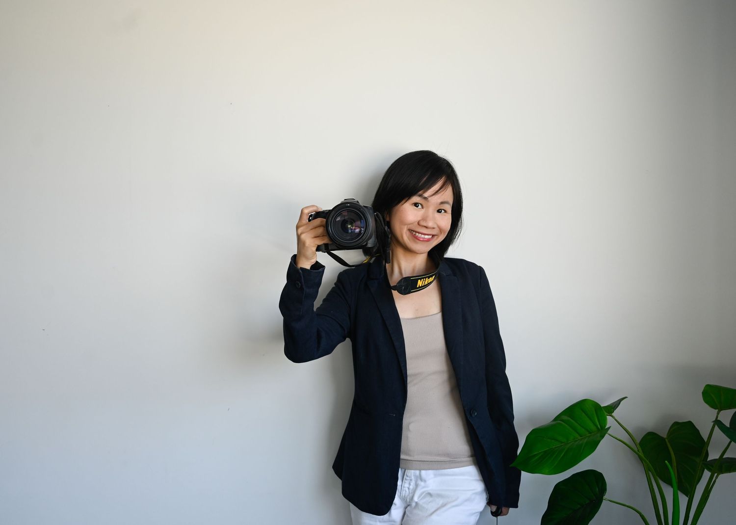 Smiling Asian woman holding a camera in a beige blazer, standing against a white wall with a green plant nearby.