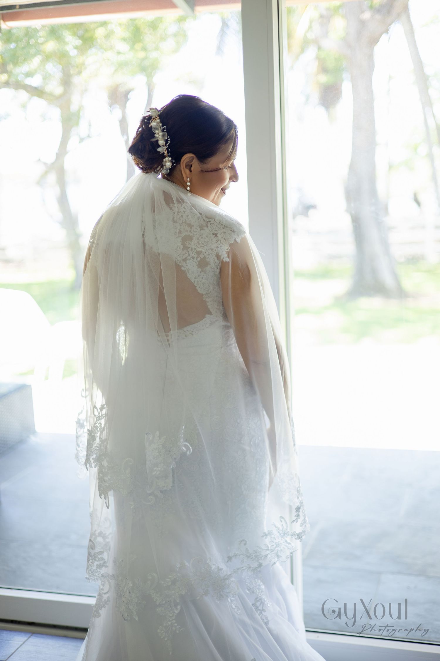 Bride in lace wedding gown and veil stands by sunlit window, showing elegant back detail and floral hair accessory.