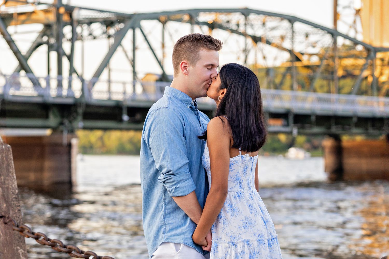 Romantic couple sharing a kiss by the river with a steel bridge in the background.