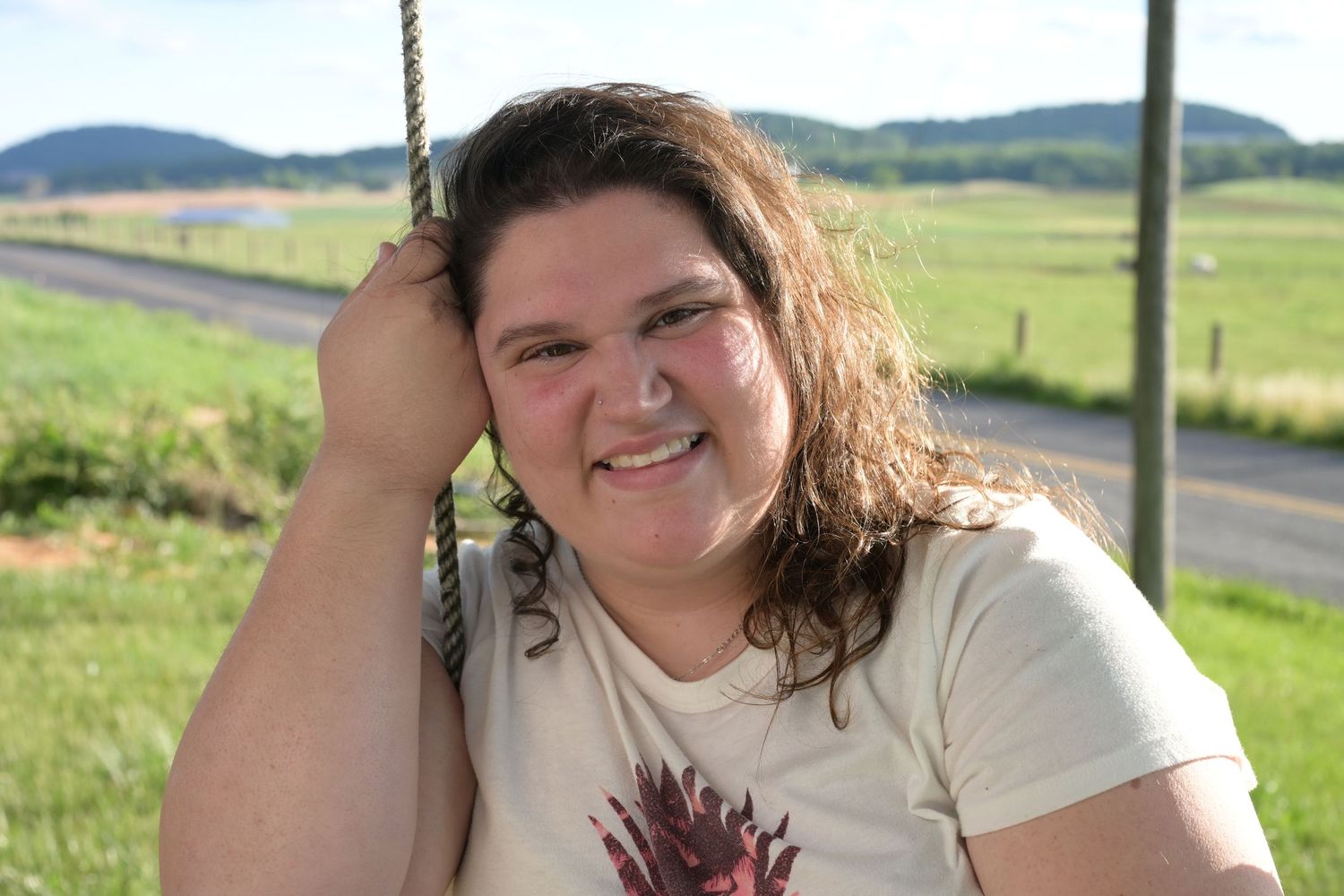Smiling woman with curly hair sitting on a rope swing outdoors with green fields and hills in the background.