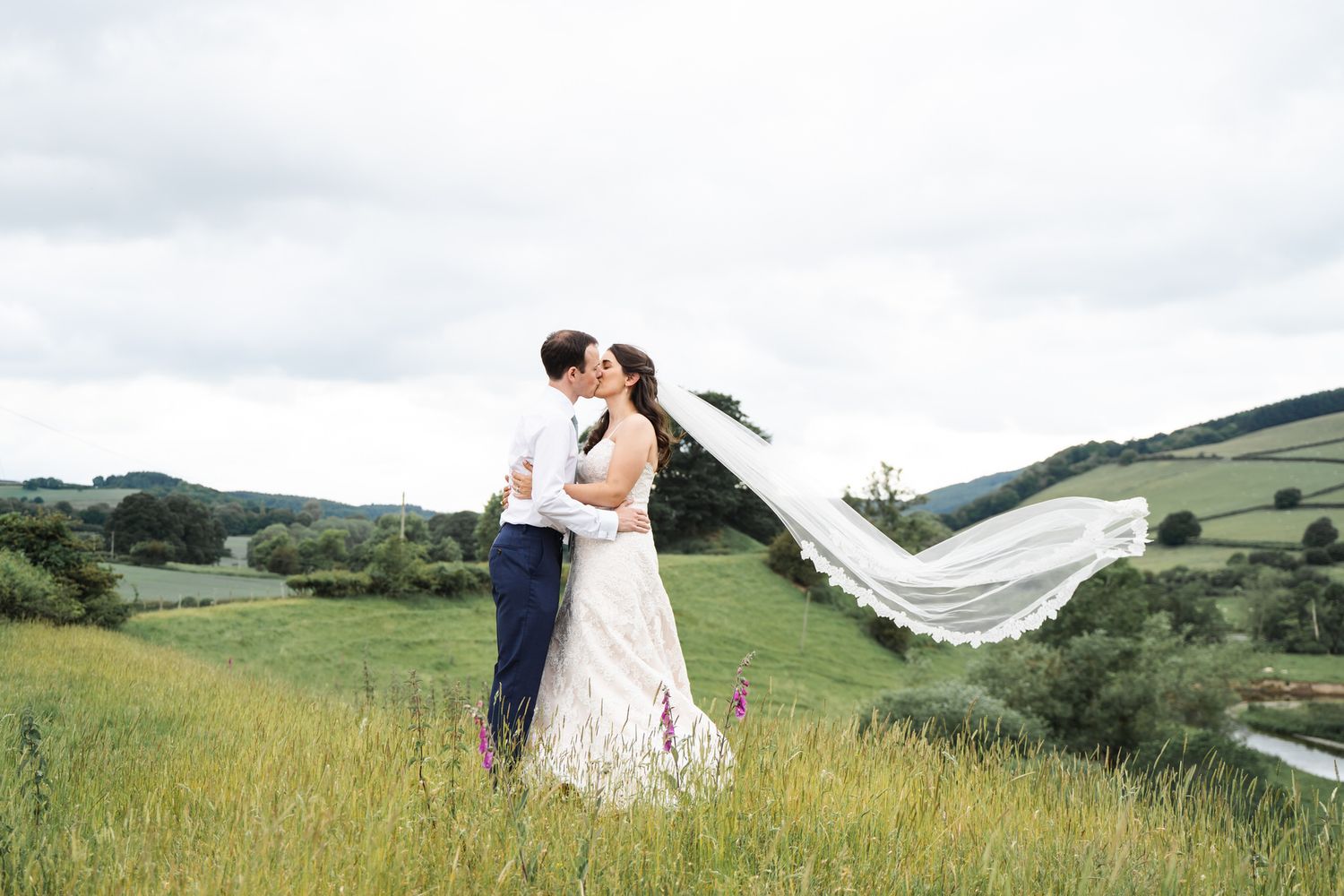 Bride and groom kissing in a green meadow with veil flowing in the wind and rolling hills in the background.