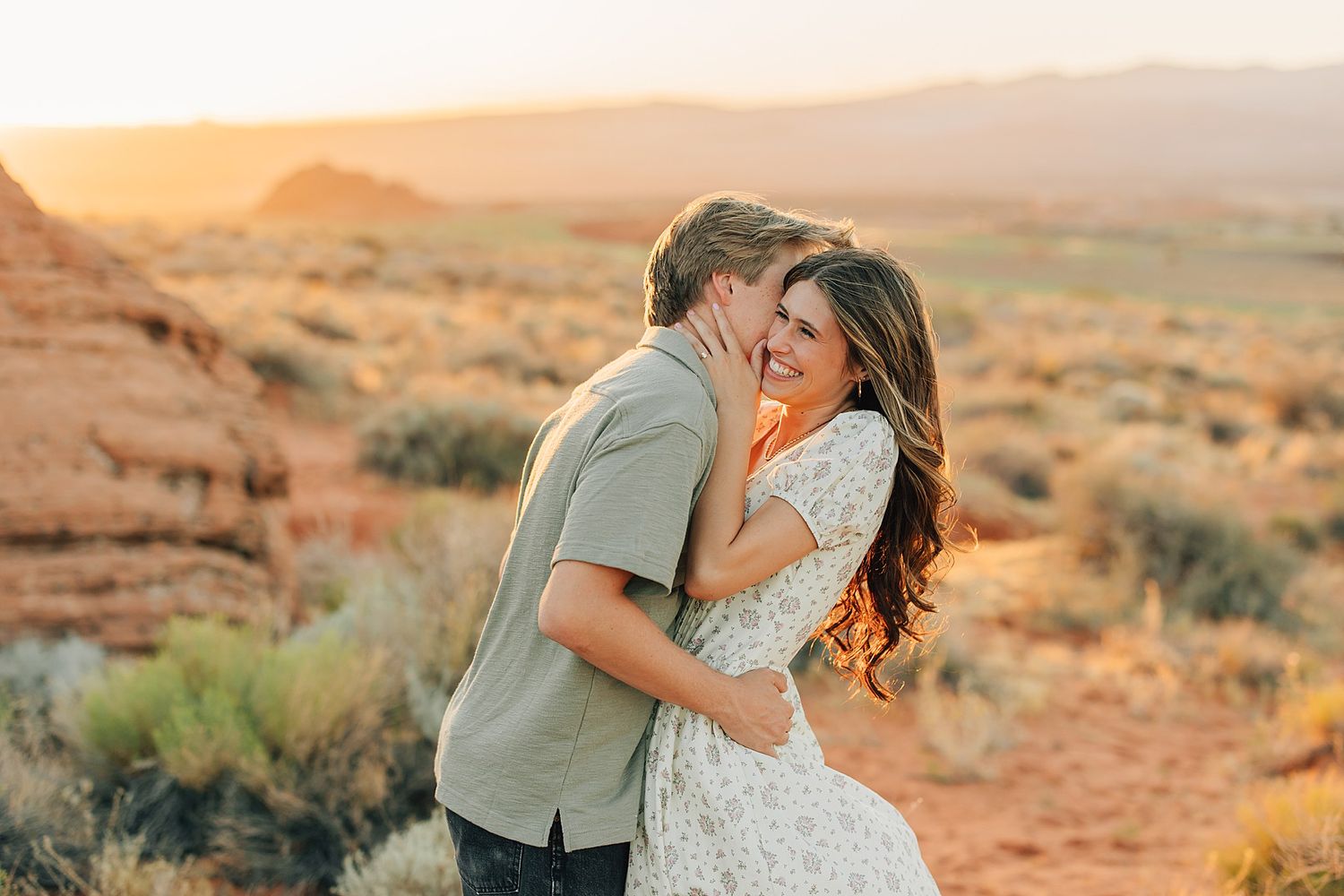 Couple sharing a romantic kiss at golden hour in a stunning red rock desert landscape with warm sunset hues.