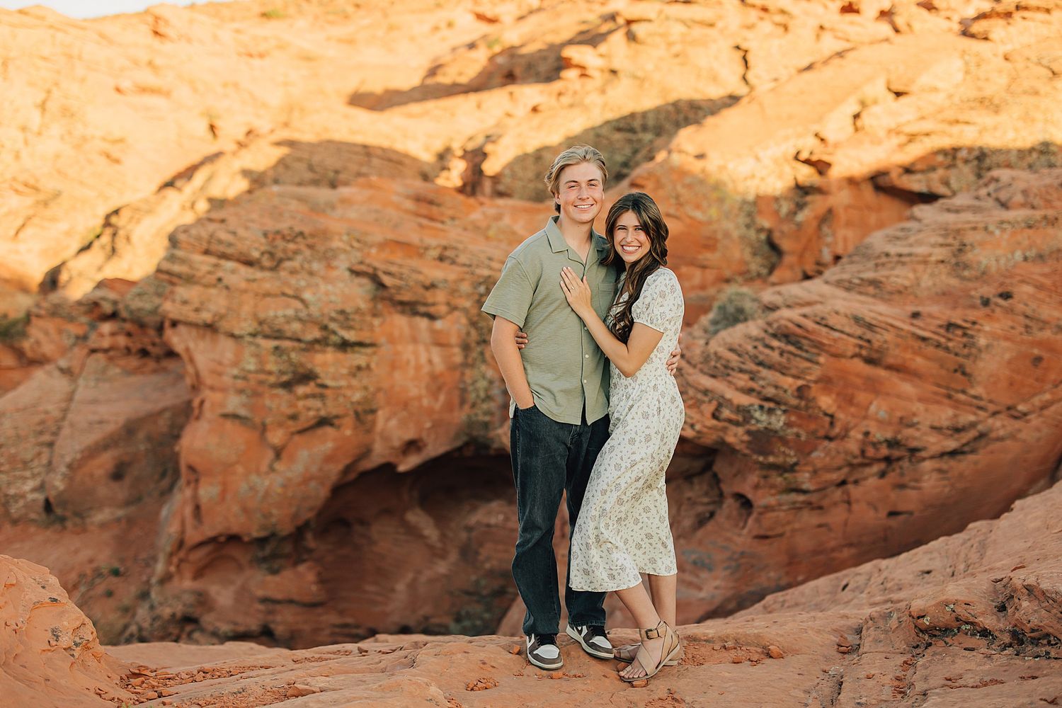 Couple embracing and smiling in front of red rock canyon formations during golden hour.