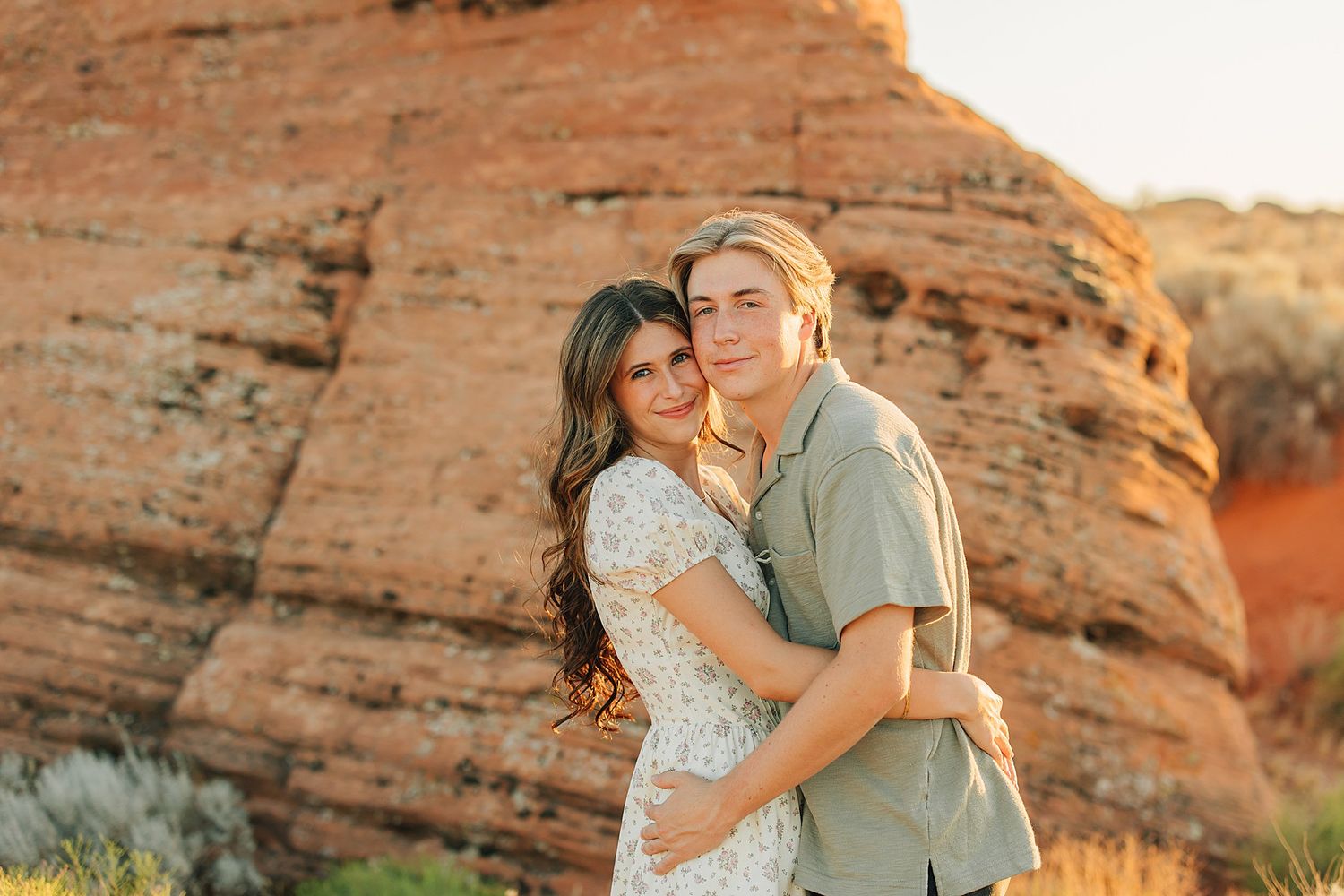 Couple embracing in front of red rock formations during golden hour in a scenic desert landscape.