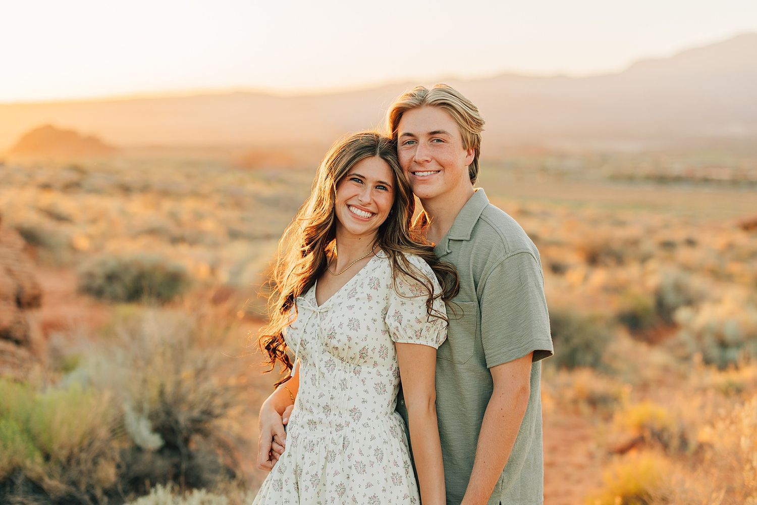 Couple smiling together at golden hour in a desert landscape with mountains in the background.