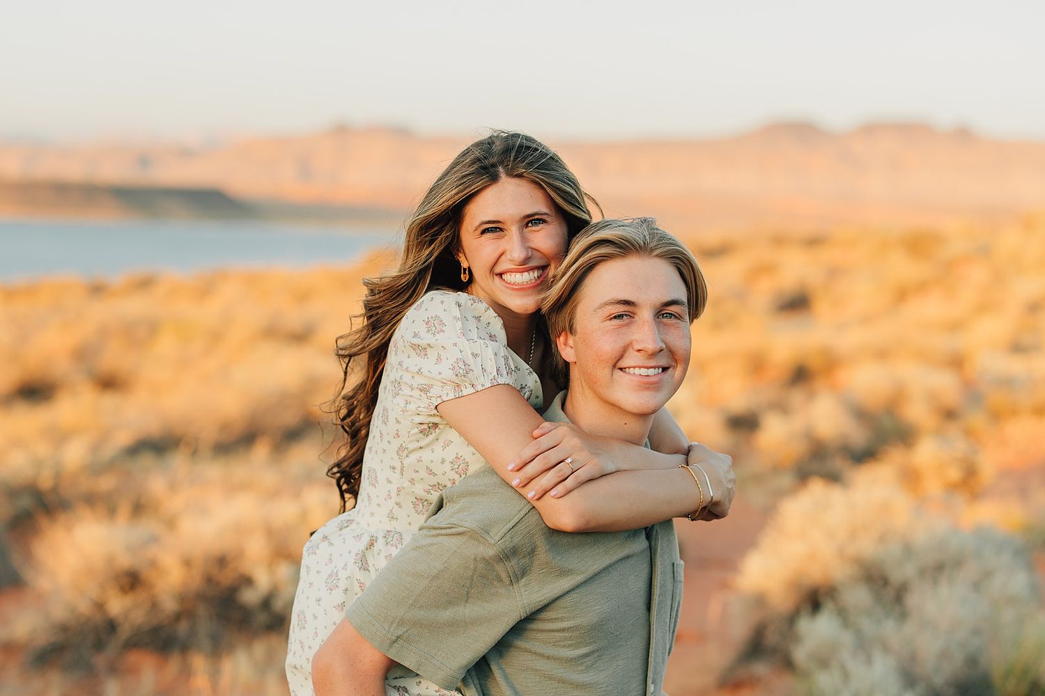 Happy couple embraces during golden hour in desert, woman hugging man from behind, smiling warmly.