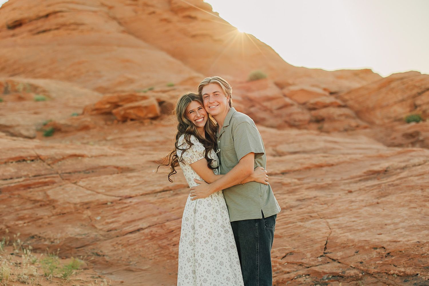 Couple embracing at sunset in front of red rock formations in a desert landscape.
