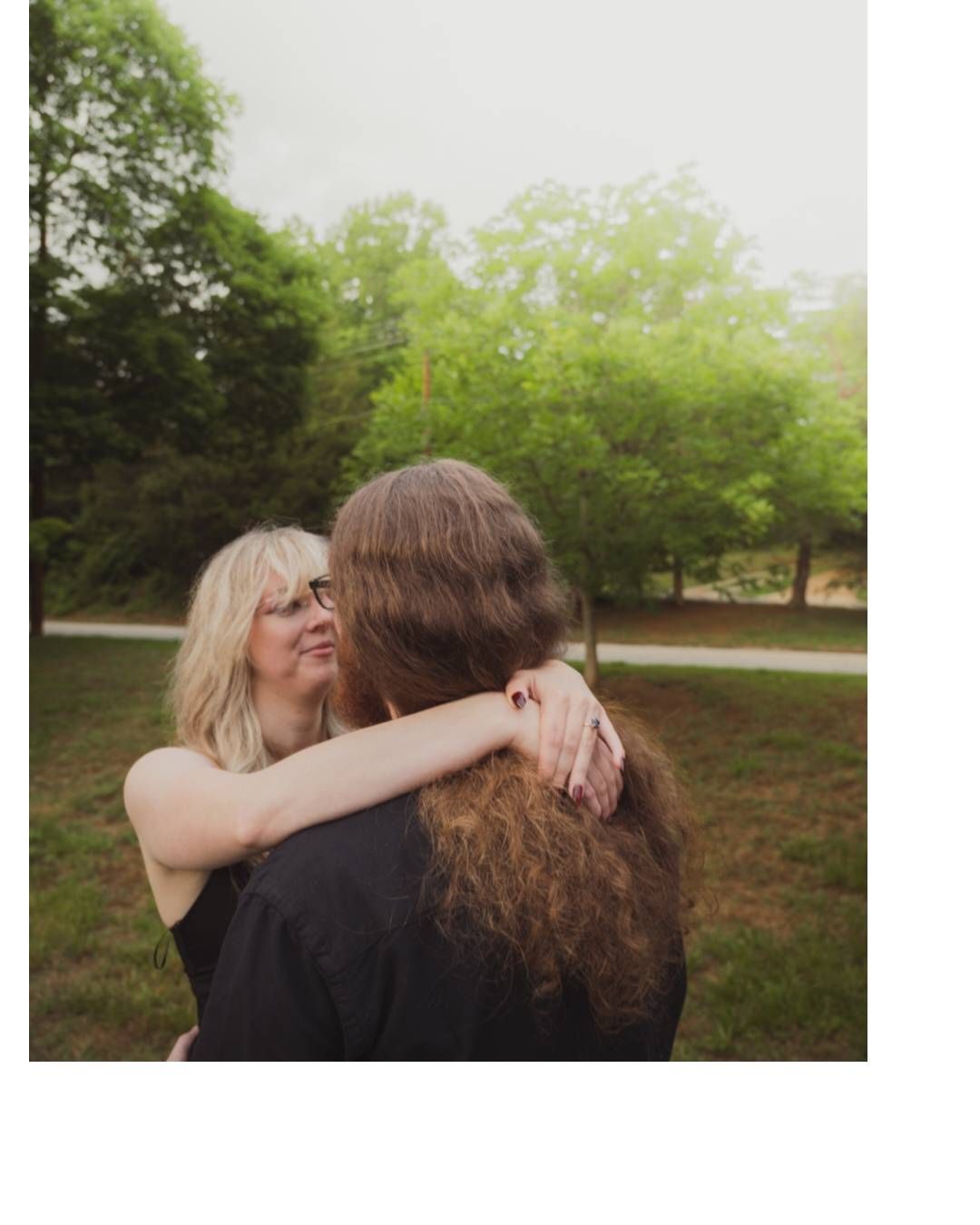 Two people sharing a warm embrace outdoors in a lush green park, with a blonde woman smiling over her partner's shoulder.