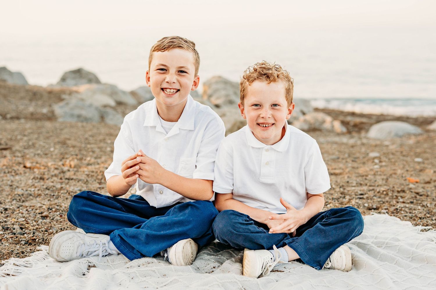 Two smiling boys in white shirts and navy pants sitting cross-legged on a blanket at a rocky beach.