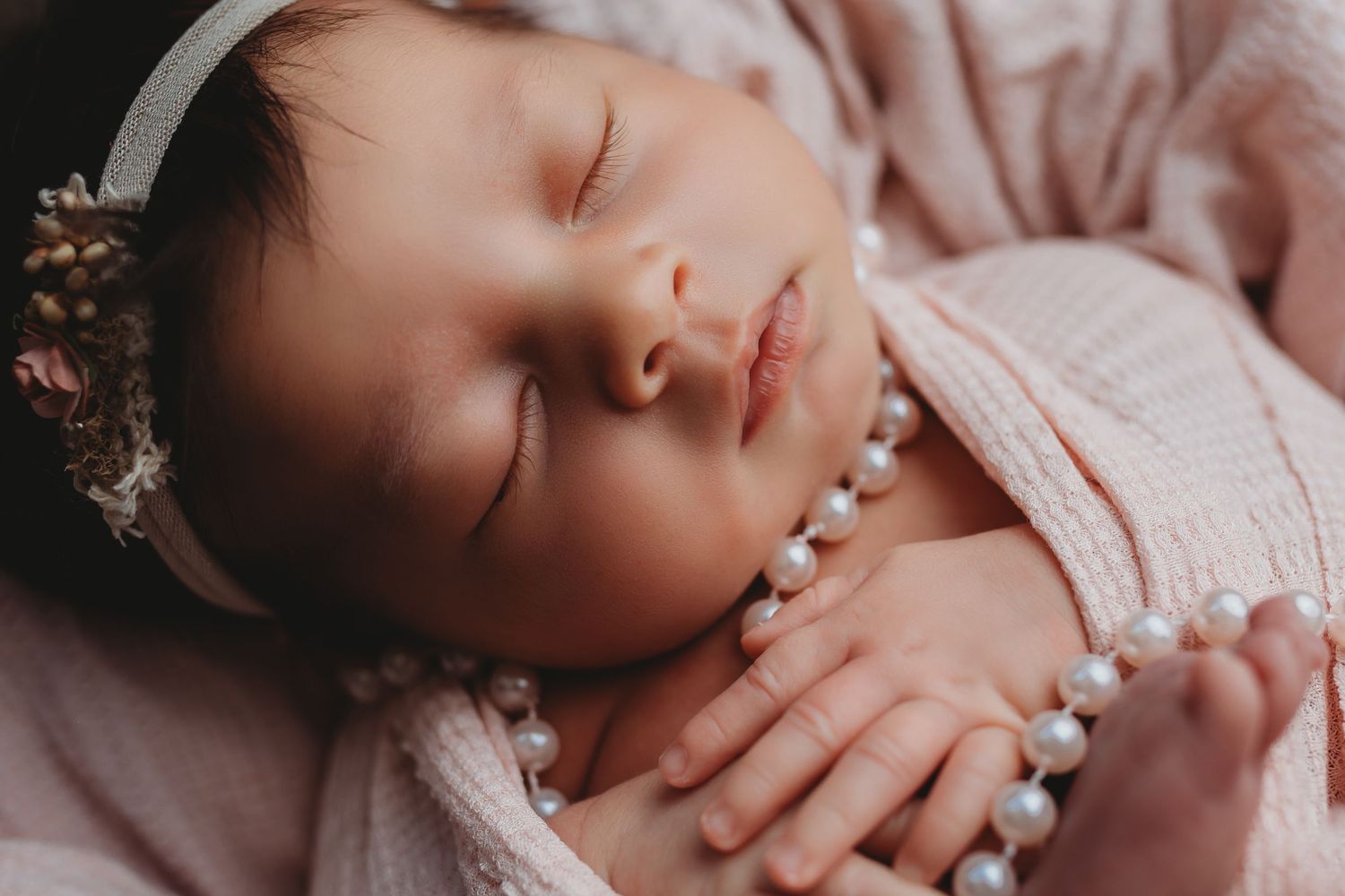 Sleeping newborn baby girl wearing pearl necklace and floral headband wrapped in soft pink fabric.