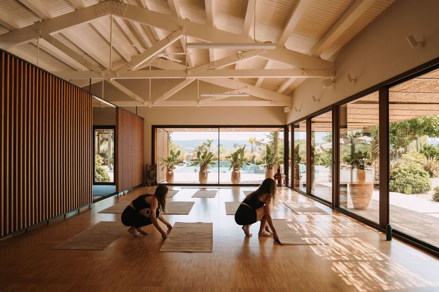 Two people practicing yoga in a sunlit studio with wooden floors, vaulted ceilings, and large glass doors overlooking a garden.