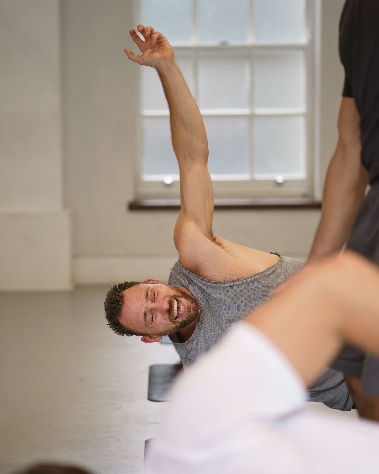 Man in gray shirt performing a yoga side stretch pose in a bright studio, smiling with arm raised.
