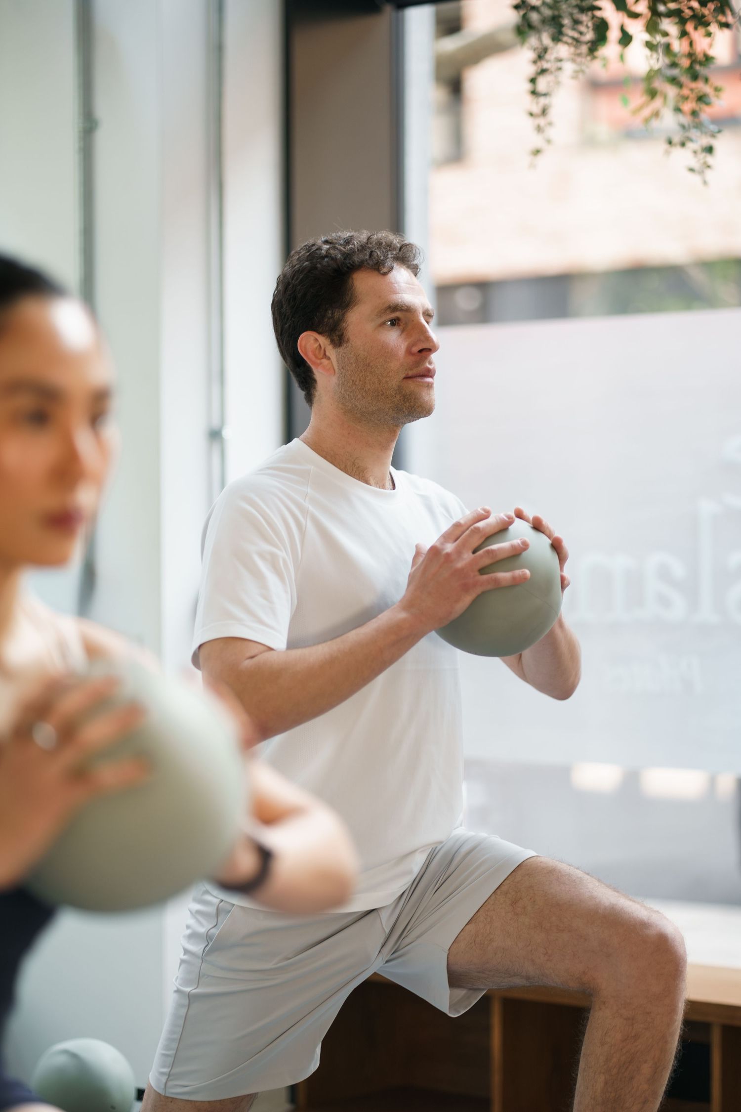 Man in white t-shirt doing lunges with medicine ball during group fitness class in modern gym.