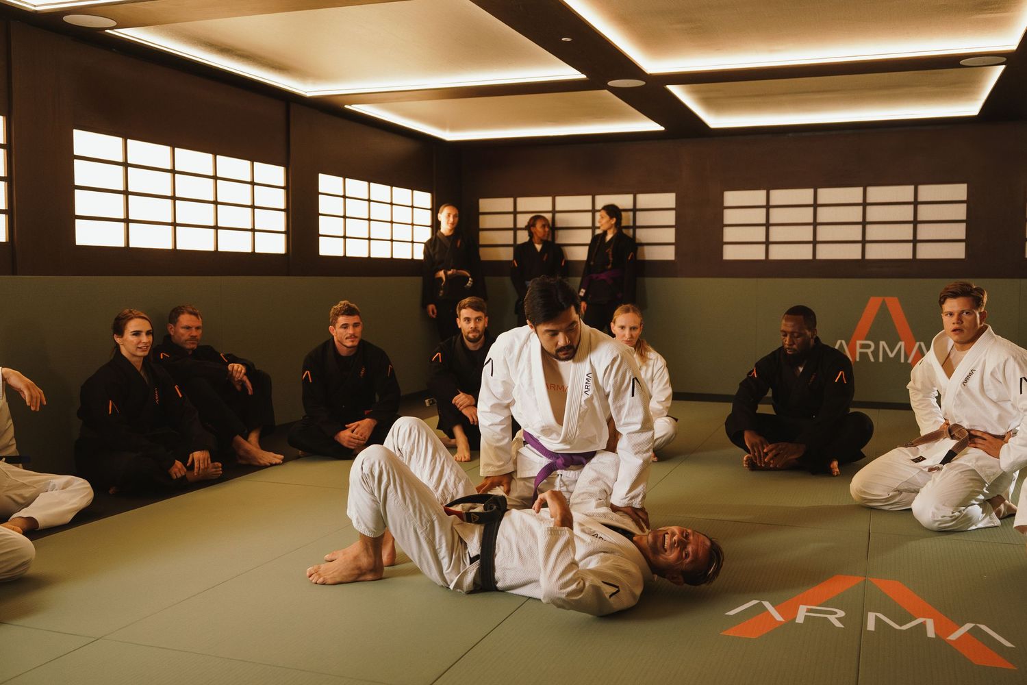 BJJ class at Arma gym with students watching a white belt grappling demonstration on the mat.