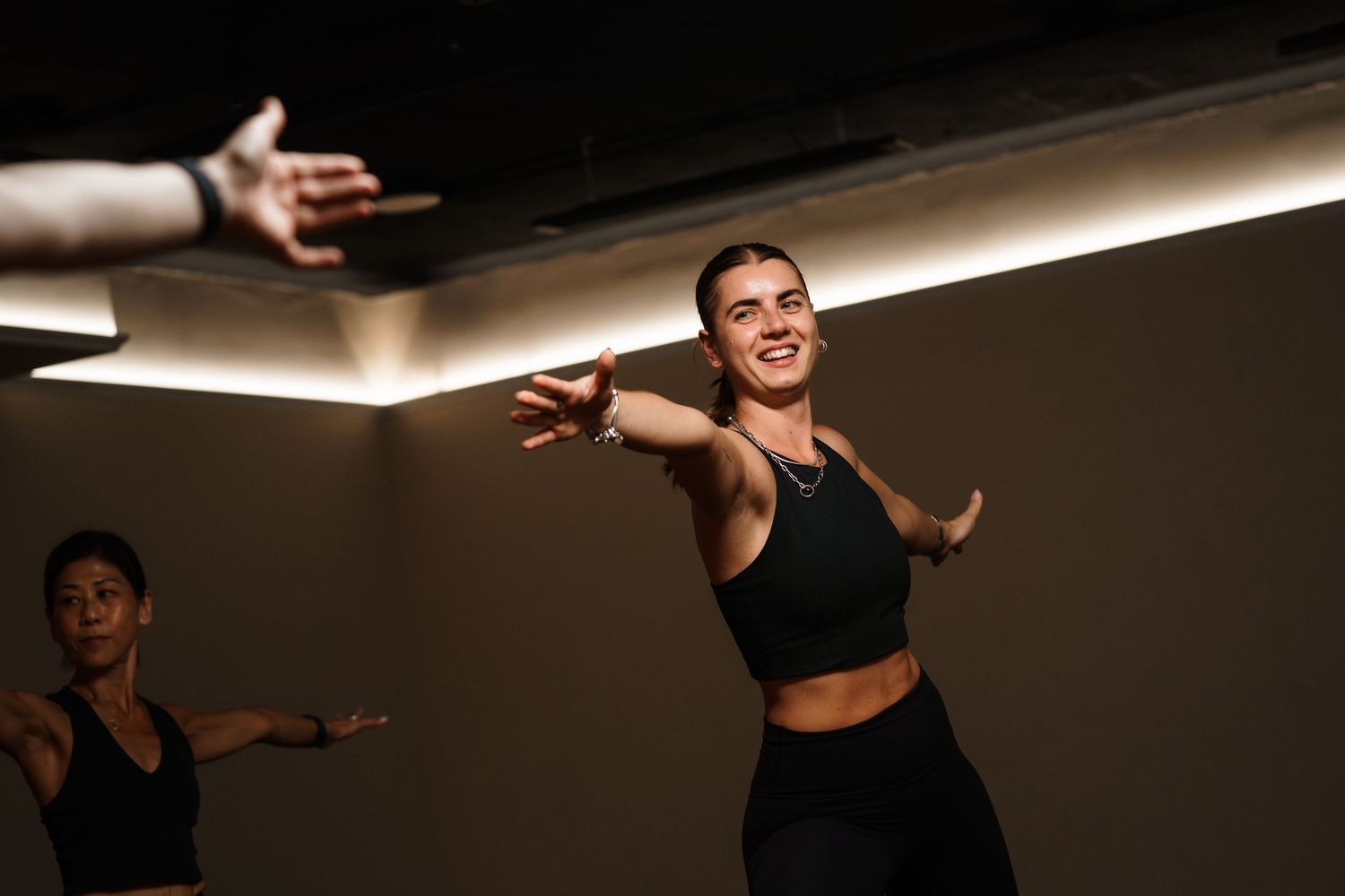 Woman in black athletic wear smiling and stretching arms during a dance fitness class in a dark studio.
