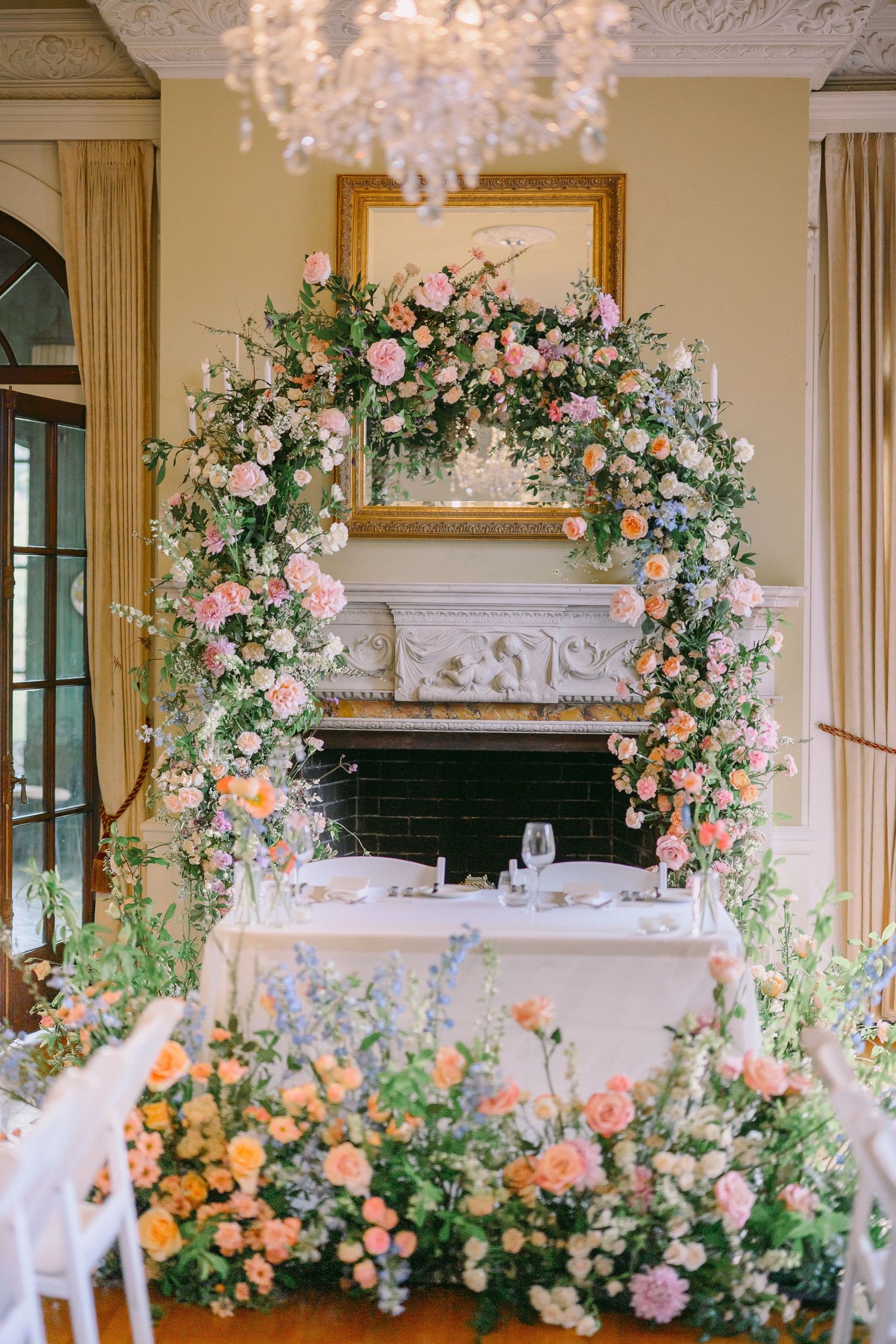 Elegant wedding sweetheart table with floral arch of pink and peach roses over a white fireplace mantel.