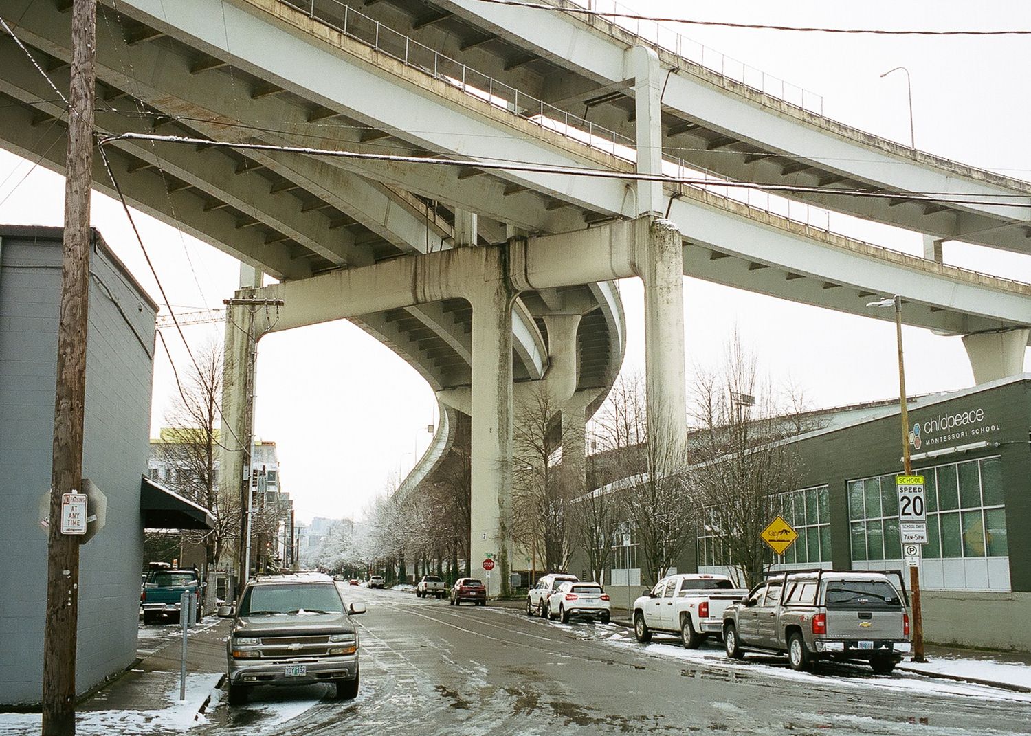 Snow-dusted urban street beneath massive concrete highway overpass with parked cars and utility poles in winter.