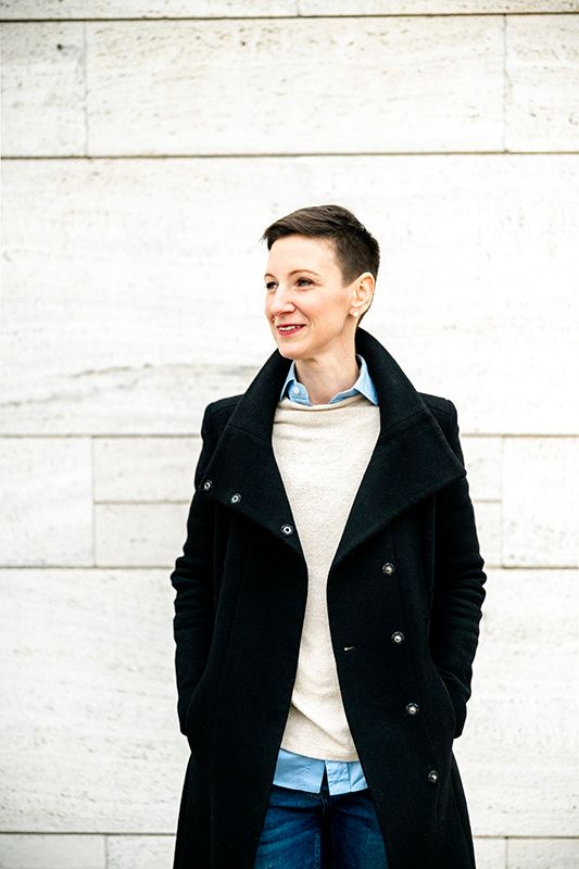 Portrait of a person in a black peacoat against a white stone wall. Eva Neuman