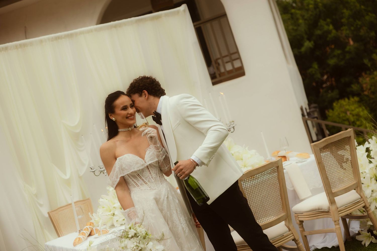 Bride in white gown and groom in white suit share a romantic moment at an elegant outdoor wedding ceremony.