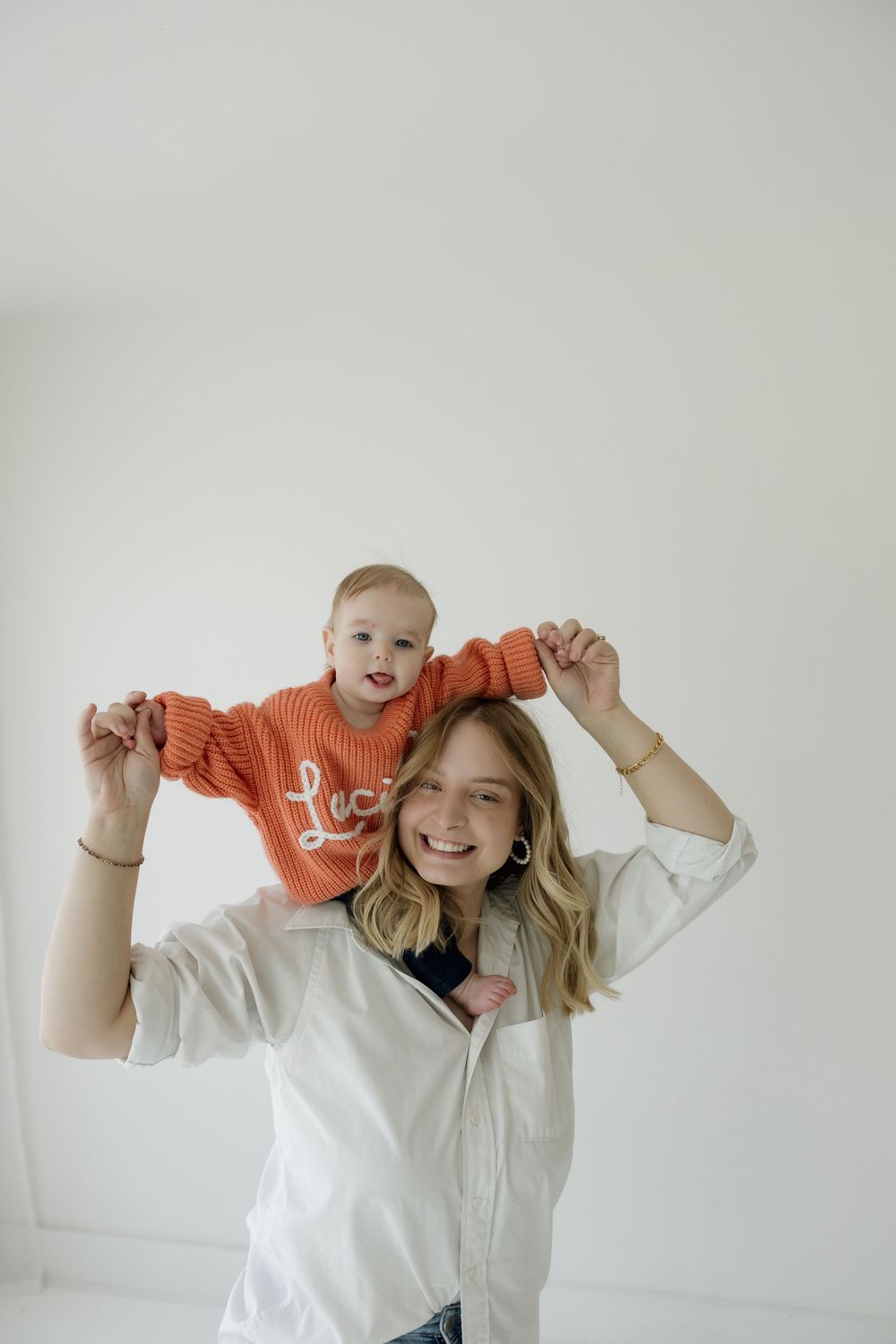 Smiling mother holding baby in orange Lucille sweater on her shoulders in bright white room.