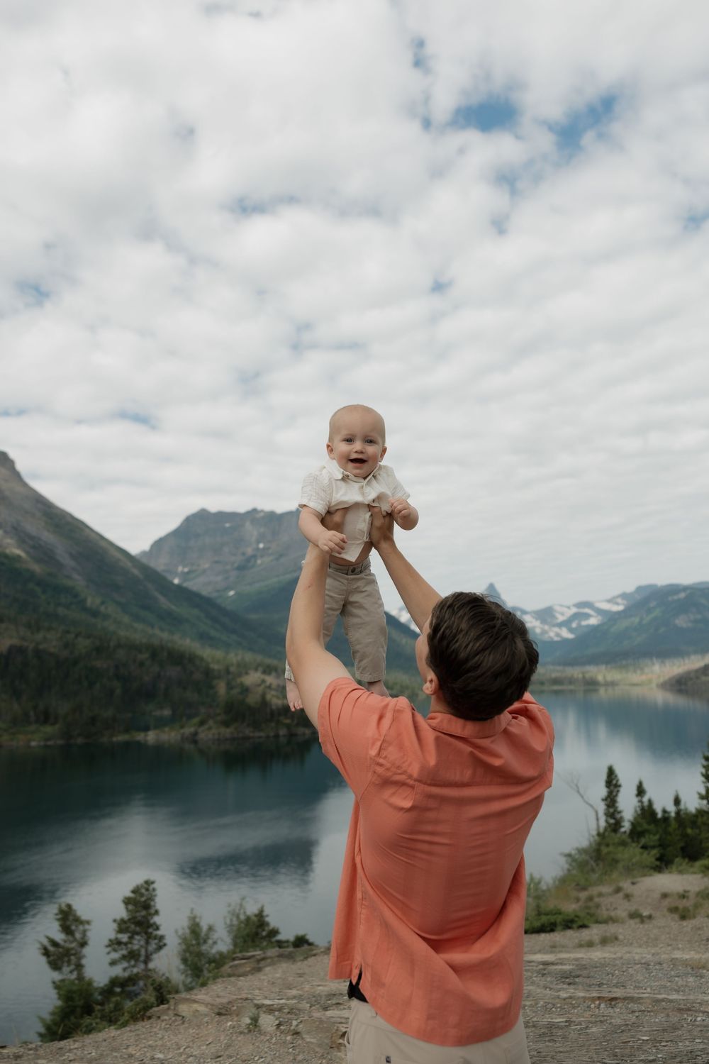 Father lifts smiling baby overhead with stunning mountain lake backdrop under cloudy sky.