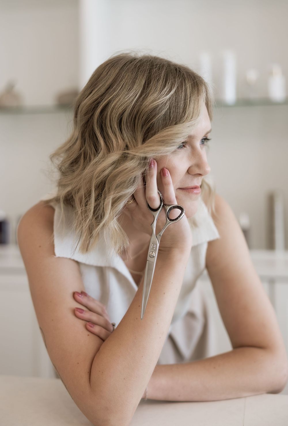 A blonde hairstylist holds scissors while sitting thoughtfully in a bright salon.