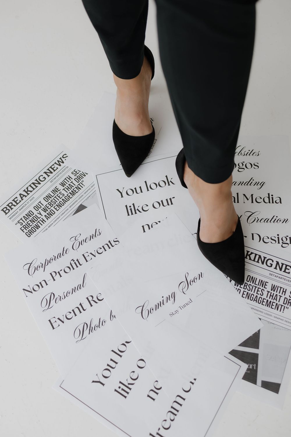 Woman in black pants and heels standing over scattered business branding printouts on white floor.