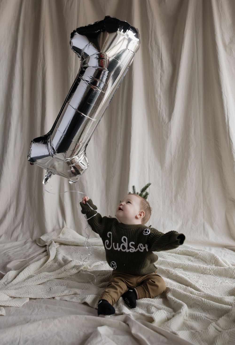 Baby boy named Judson sits on white fabric holding a silver number 1 balloon on his first birthday.