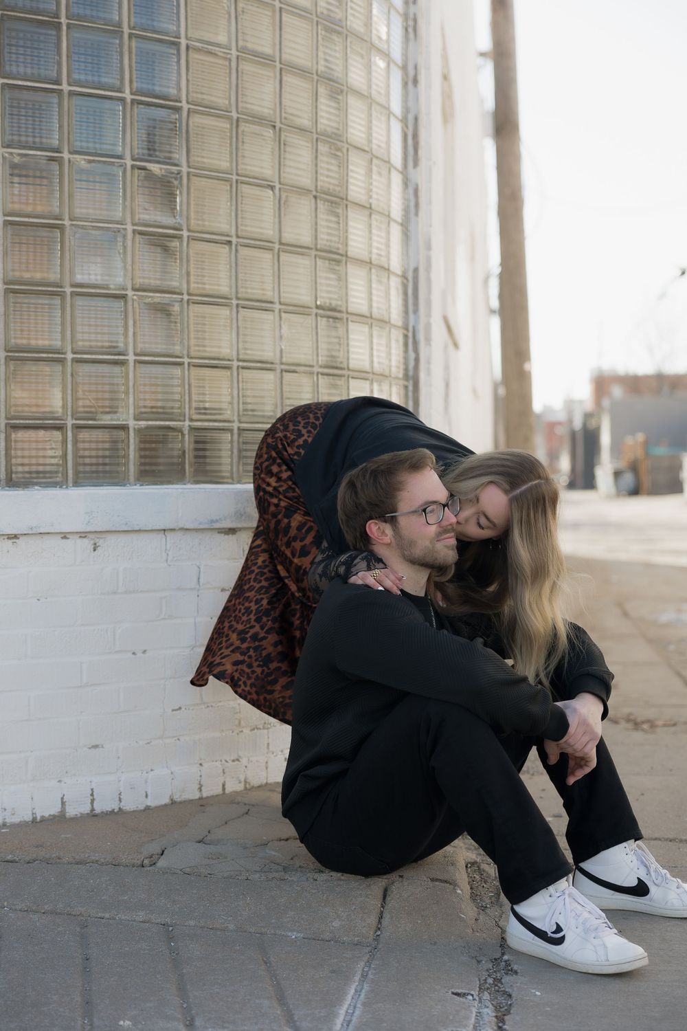 Affectionate couple sitting on urban sidewalk, woman leaning over mans shoulders near glass block building wall.