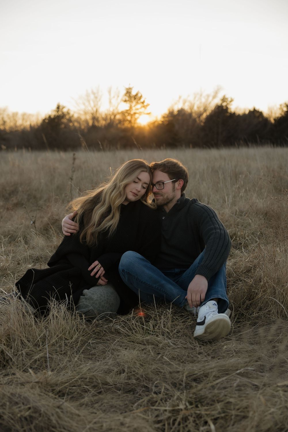 Romantic couple sitting together in dry grass field at golden hour sunset, leaning close together.