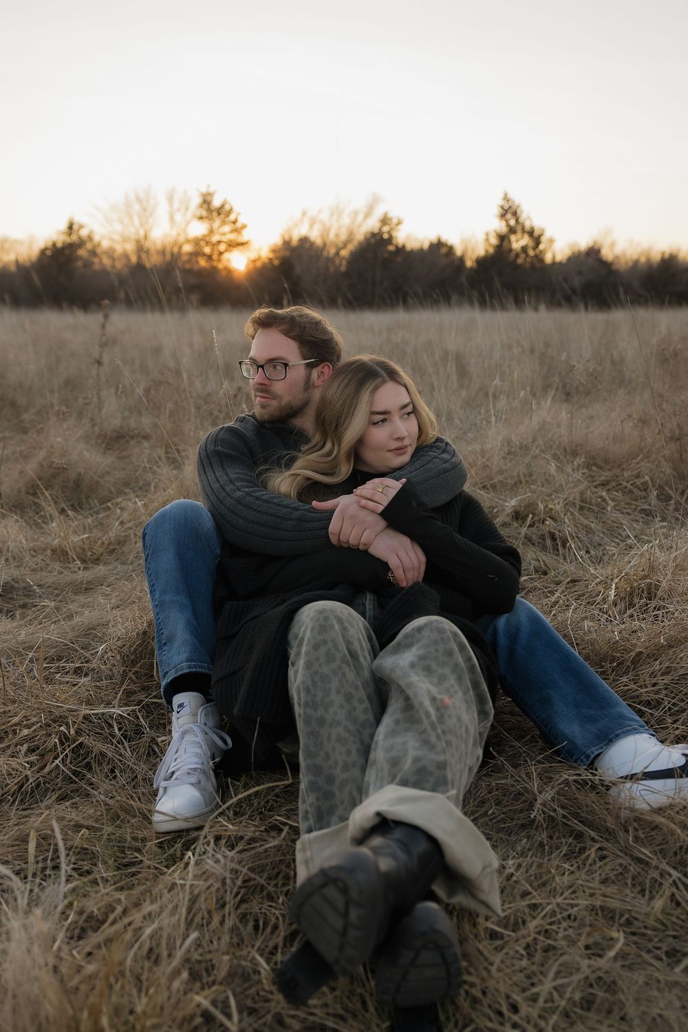 Couple sitting in dry autumn field at sunset, woman resting head on mans shoulder wearing cozy boots.