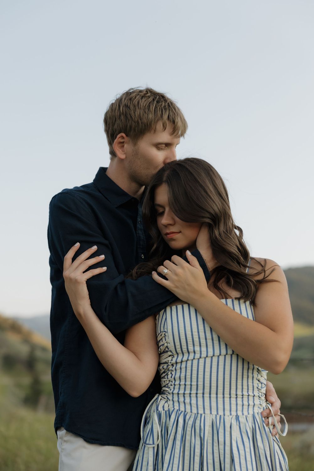 A couple embraces outdoors, man in navy shirt kisses woman's head as she leans into him wearing a striped dress.