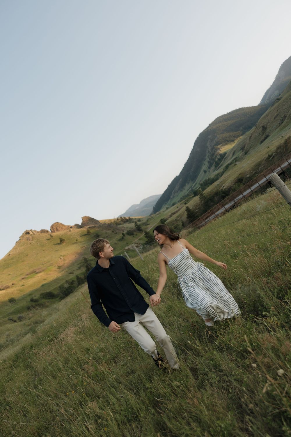 A couple holds hands while walking through a lush green mountain meadow, the woman in a flowing dress smiling joyfully.